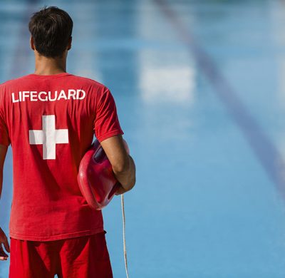 Rear view of male lifeguard with emergency equipment in red uniform watching swimming pool.